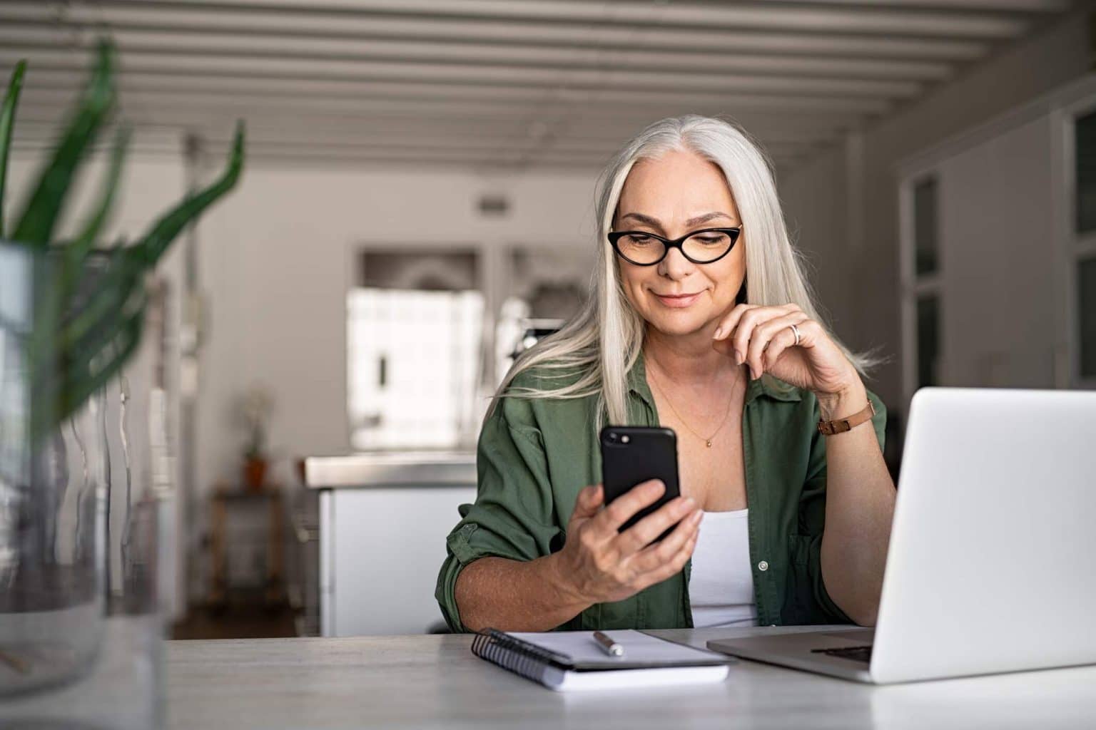 Mature woman looking at phone while in front of laptop